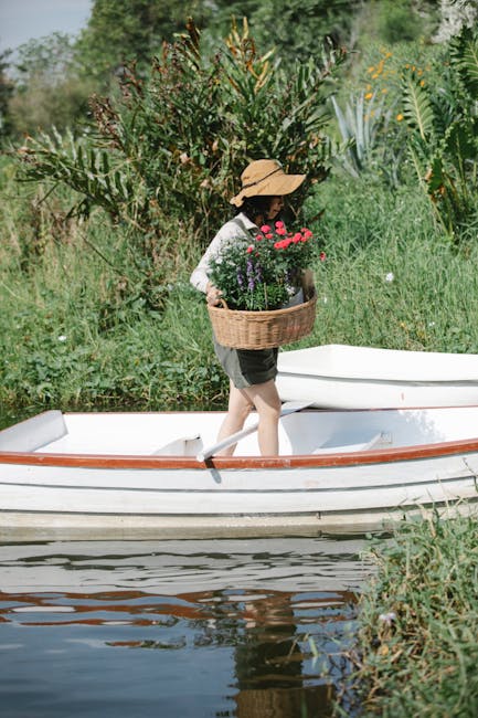 A person wearing a wide-brimmed straw hat, a long-sleeved patterned shirt, and shorts walks onto a small white boat with wooden trim, carrying a wicker basket filled with vibrant pink, purple, and red flowers. The boat is positioned on a calm water surface, reflecting the boat's underside and nearby foliage. Surrounding the scene are lush green plants, tall grasses, and shrubs, with some plants featuring yellow flowers. In the background, a densely planted garden area with various trees and bushes provides a natural, slightly wild setting. The scene is illuminated with natural daylight, highlighting the textures of the wet soil, trimmed grass, and leafy plants. This outdoor scene illustrates careful garden management and landscaping work supported by Gardeners Barnes, emphasizing their expertise in maintaining riverside gardens and promoting visual harmony between planted borders and water features.