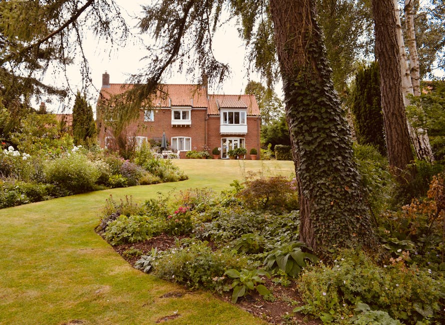A lush, well-maintained garden featuring a variety of plants, shrubs, and flowers arranged along a gently curved pathway with neatly trimmed grass. In the foreground, large mature trees with textured bark and leafy branches provide shade and structure to the landscape. The garden beds contain flowering plants with colorful blooms and dense foliage, showcasing different textures and heights. A broad lawn area extends toward a red-brick period house in the background, with white-framed windows and a small patio area. The scene captures a calm, organized outdoor space with evidence of garden maintenance such as pruned bushes and tidy borders, ideal for landscaping work or lawn care, with natural light evenly illuminating the scene. The image reflects professional gardening care, possibly undertaken by Gardeners Barnes, to enhance the aesthetic and functionality of a traditional London home’s garden.